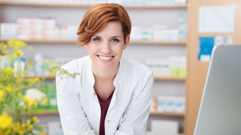 Eine lächelnde Frau mit kurzen roten Haaren trägt einen weißen Laborkittel. Sie blickt direkt in die Kamera. Im unscharfen Hintergrund sind Apothekenregale mit Medikamenten und Produkten zu erkennen.