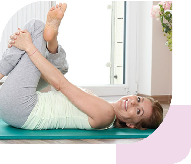 Eine Frau liegt auf dem Rücken auf einer Yogamatte. Ihre Beine sind angehoben und ihre Knie sind im rechten Winkel gebeugt. Ihre Arme sind ebenfalls leicht angehoben. Im Hintergrund sind große Fenster und eine Vase mit Blumen zu sehen.