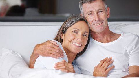 Ein älteres Pärchen liegt kuschelnd im Bett und lächelt in die Kamera