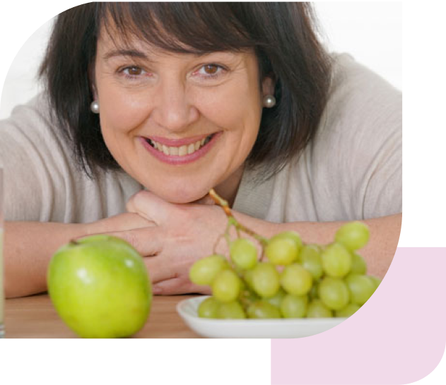 Eine Frau lächelt in die Kamera und lehnt auf einem Tisch. Im Vordergrund liegt ein Apfel und eine Schale mit grünen Weintrauben.